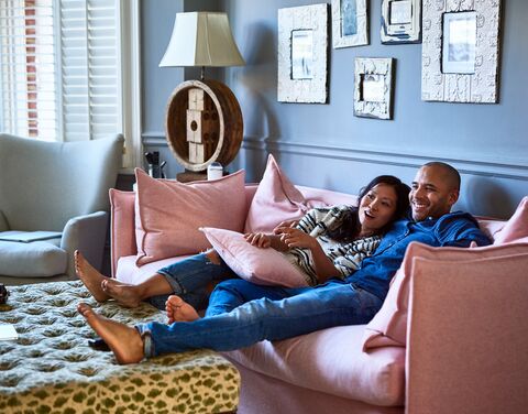 A smiling couple relaxes on a pink sofa in a cozy living room, surrounded by cushions and decorations.