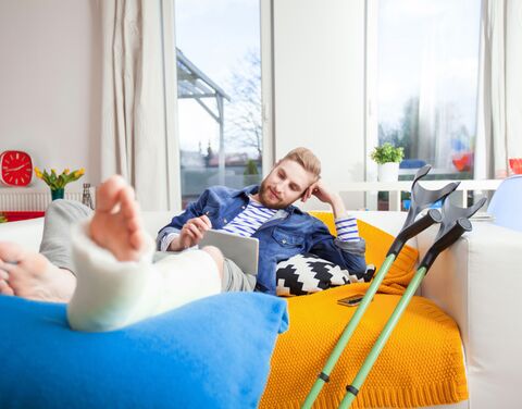 A man with a leg in plaster lies on a sofa, surrounded by crutches and cushions, using a tablet in a bright, cozy living room.