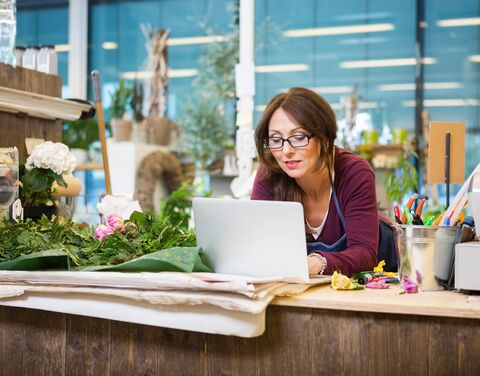 Eine Floristin mit Brille arbeitet an einem Laptop in ihrem Blumenladen, umgeben von Blumen, Pflanzen und Arbeitsutensilien.