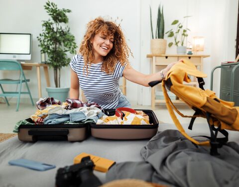 Une femme souriante aux cheveux bouclés prépare une valise dans une pièce lumineuse, entourée de plantes et de meubles modernes.