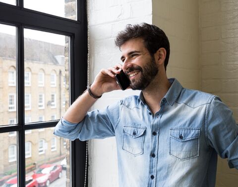 Un homme souriant, vêtu d'une chemise en jean bleu clair, téléphone à côté d'une grande fenêtre à travers laquelle on aperçoit une façade en briques et des voitures garées.