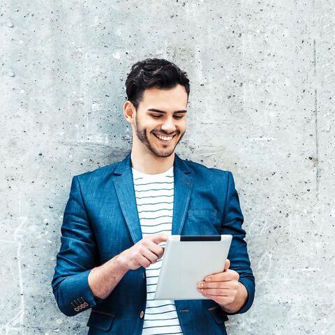 A man in a blue blazer and striped T-shirt smiles while using a tablet in front of a gray concrete wall.