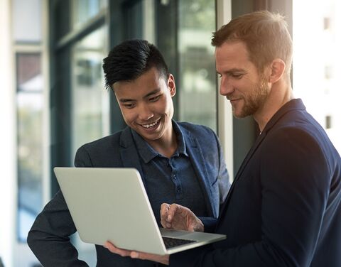 Two men in business attire stand in a modern office, smiling and looking at a laptop held by one of them.