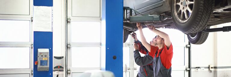 Two mechanics in work clothes repair a car that is raised on a lifting platform in a bright and modern workshop.