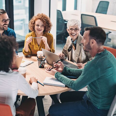 Un groupe de cinq personnes est assis autour d'une table ronde dans un bureau moderne et discute joyeusement tout en prenant des notes et en utilisant des ordinateurs portables.