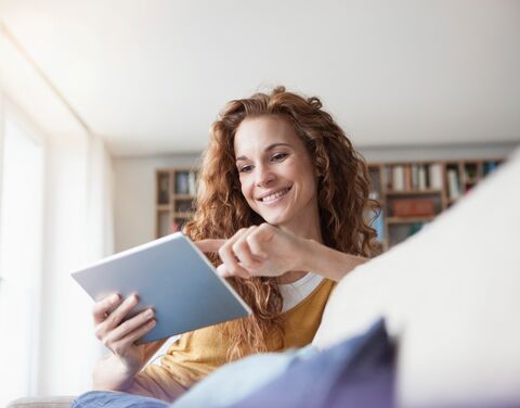 Eine lächelnde Frau mit lockigem Haar sitzt auf einem Sofa und benutzt ein Tablet, umgeben von Büchern in einem hellen Raum.