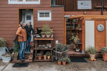 Ein älteres Paar steht vor einem kleinen, orangefarbenen Haus mit vielen Pflanzen in Töpfen und unterhält sich freundlich.