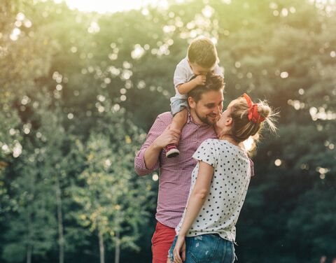 Una coppia sorridente si trova all'aperto davanti a degli alberi, l'uomo porta un bambino sulle spalle mentre la donna gli dà un bacio.