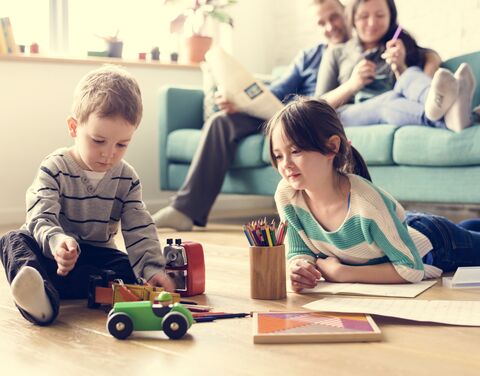 Two children play on the floor with toys and crayons, while a couple sits relaxed on a sofa in the background.