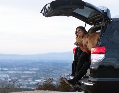 Eine Frau sitzt mit einem Hund im geöffneten Kofferraum eines Autos und geniesst den Ausblick auf eine weite Landschaft im Hintergrund.